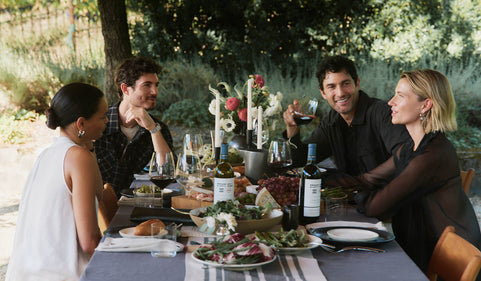 Group of people sitting around a table outdoors, enjoying a meal and drinks.