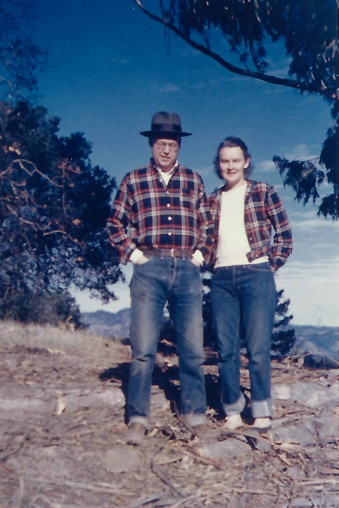 Two people wearing plaid jackets standing outdoors with trees and mountains in the background.