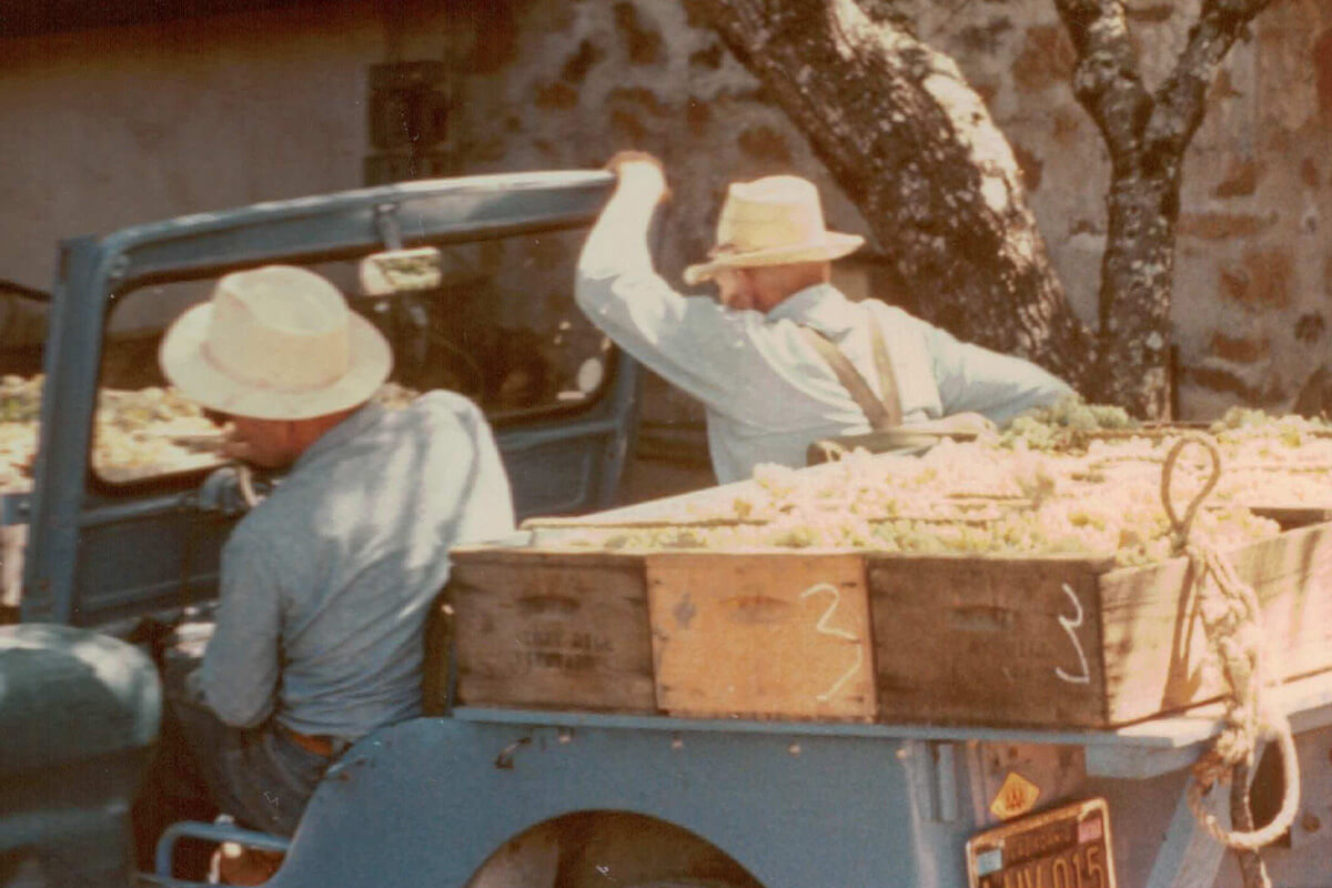Two men in hats loading produce into a truck with a stone building in the background