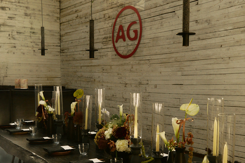 Dining area with floral arrangements and candles on a wooden wall with 'AG' logo.