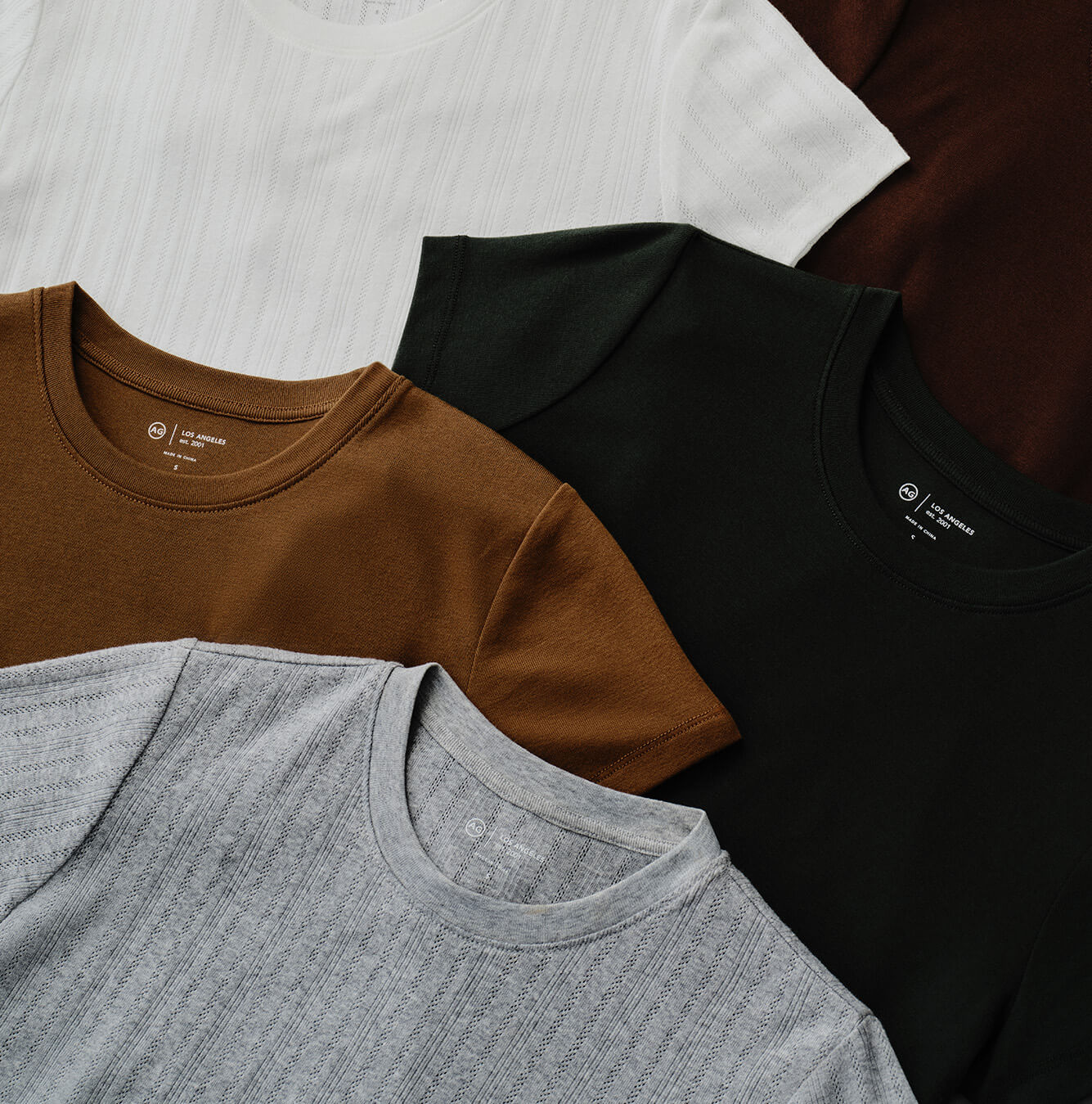 Four crewneck t-shirts arranged diagonally. The shirts are in white, brown, tan, black, and heather gray.