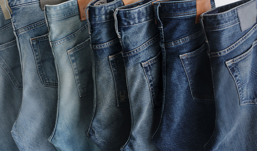 A close-up row of blue denim jeans hanging side by side, showing different washes from light blue to dark navy, with the back pockets and waistbands visible.