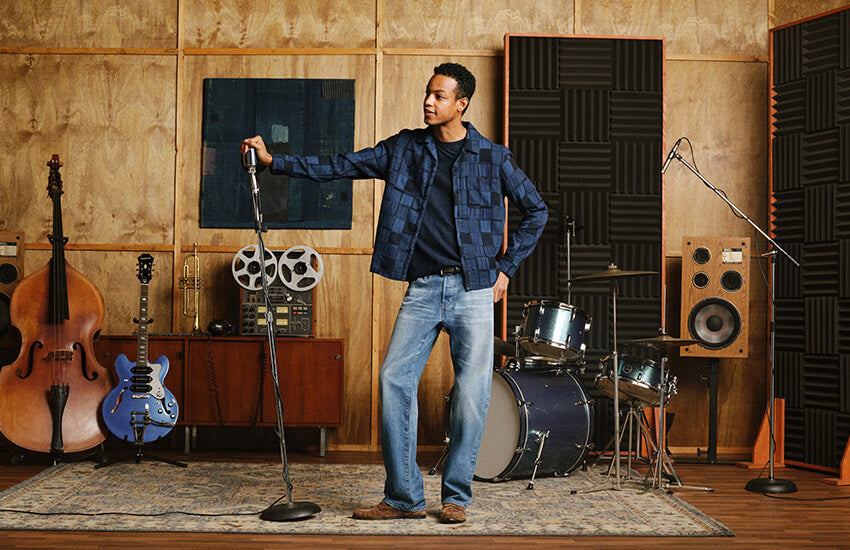 A man stands in a music studio wearing a blue plaid flannel shirt and jeans, holding a microphone stand. The room features wood-paneled walls, acoustic foam panels, a double bass, a blue electric guitar, a drum kit, and speakers.