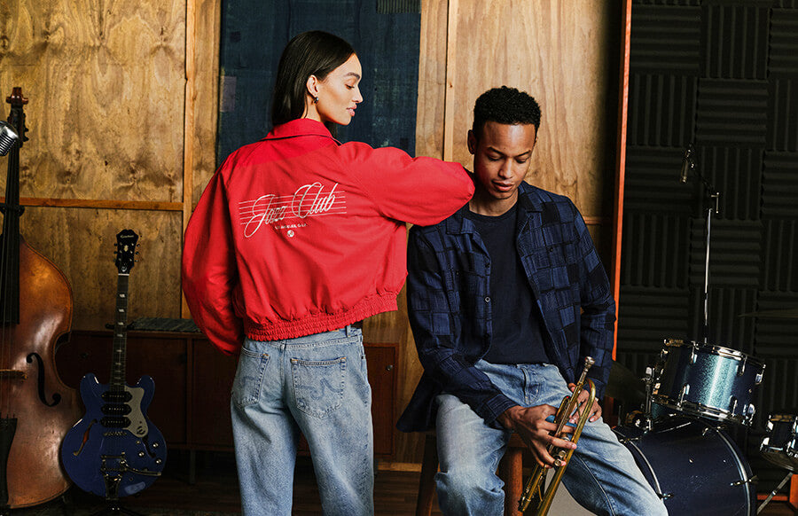 A woman in a red bomber jacket with 'Jazz Club' written on the back stands next to a man in a music studio holding a trumpet. The studio features wooden walls, acoustic panels, and musical instruments including a double bass, blue electric guitar, and drums.