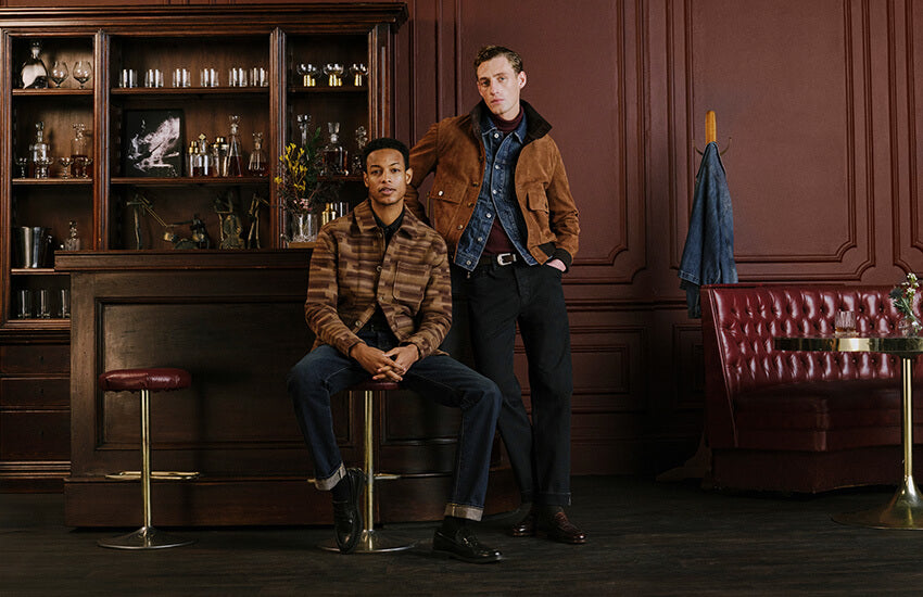 Two men modeling outerwear in a dark, elegant bar interior. One man sits on a red leather bar stool wearing a brown and black plaid jacket, blue jeans, and black boots.