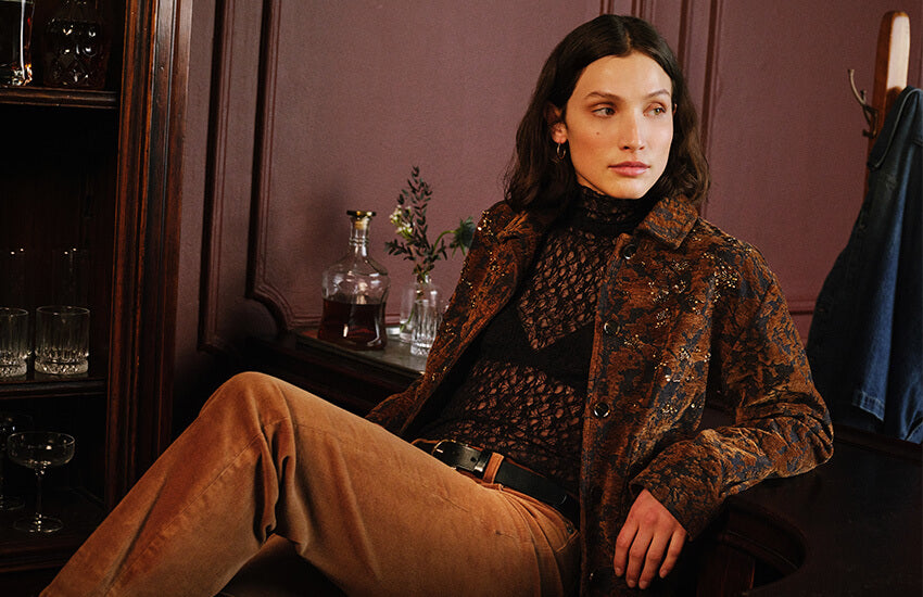 A woman seated indoors wearing a dark brown patterned jacket over a black top and tan pants, posed in a dimly lit room with wood paneling and furniture in the background.