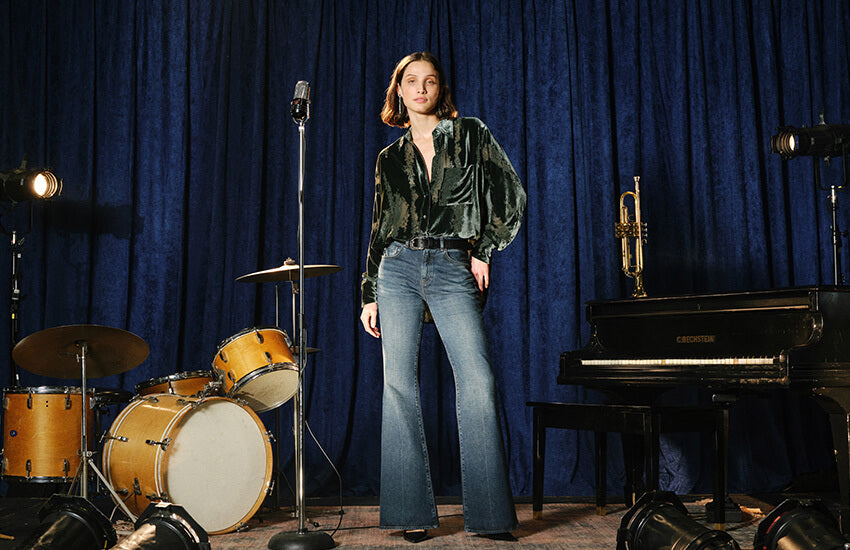 A woman stands in a studio setting with a blue curtain backdrop, wearing a dark green velvet long-sleeve top and blue flared jeans. Musical instruments, including a drum set, microphone, trumpet, and piano, are arranged around her.