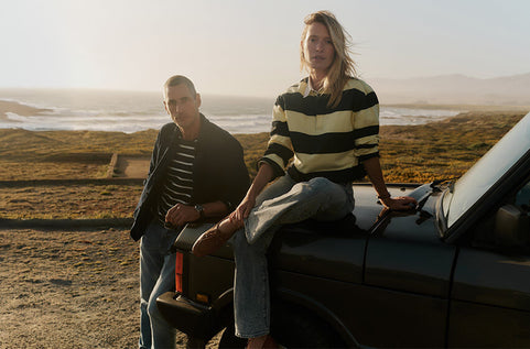 Two people sitting on a car in a desert landscape with mountains in the background.
