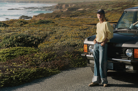 Person leaning against a vintage car by a scenic coastal road