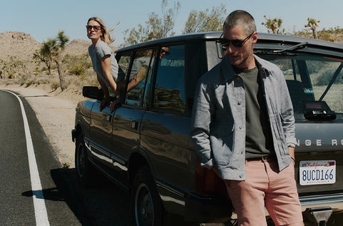 Two people standing by a car on a desert road with Joshua Tree in the background