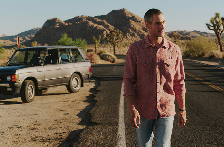 Man standing on a desert road with a vintage car, surrounded by mountains and cacti.