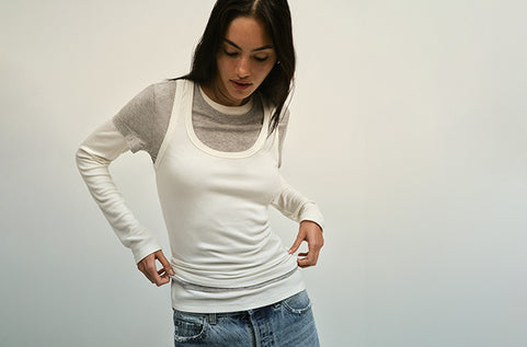 Woman wearing a white long-sleeve top over a gray tank top with blue jeans on a plain background