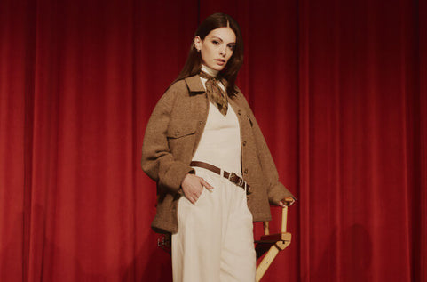 Woman in a brown jacket and white dress standing against a red curtain