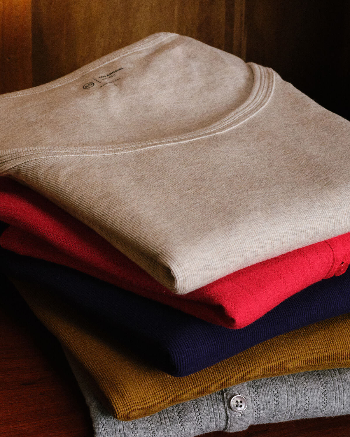 A stack of folded ribbed knit tops in multiple colors, including beige, red, navy, mustard yellow, and gray, arranged on a dark wooden surface.