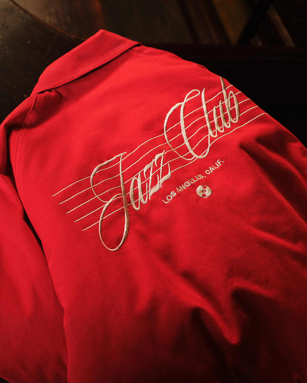A close-up of a red collared shirt or jacket with white embroidered script reading 'Jazz Club' across a musical staff, plus 'Los Angeles, Calif.' and a small circular emblem near the chest.