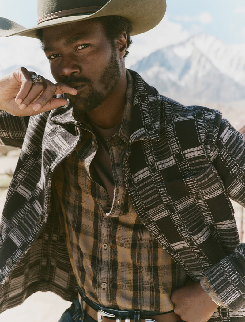 Man wearing a cowboy hat and plaid jacket with mountains in the background