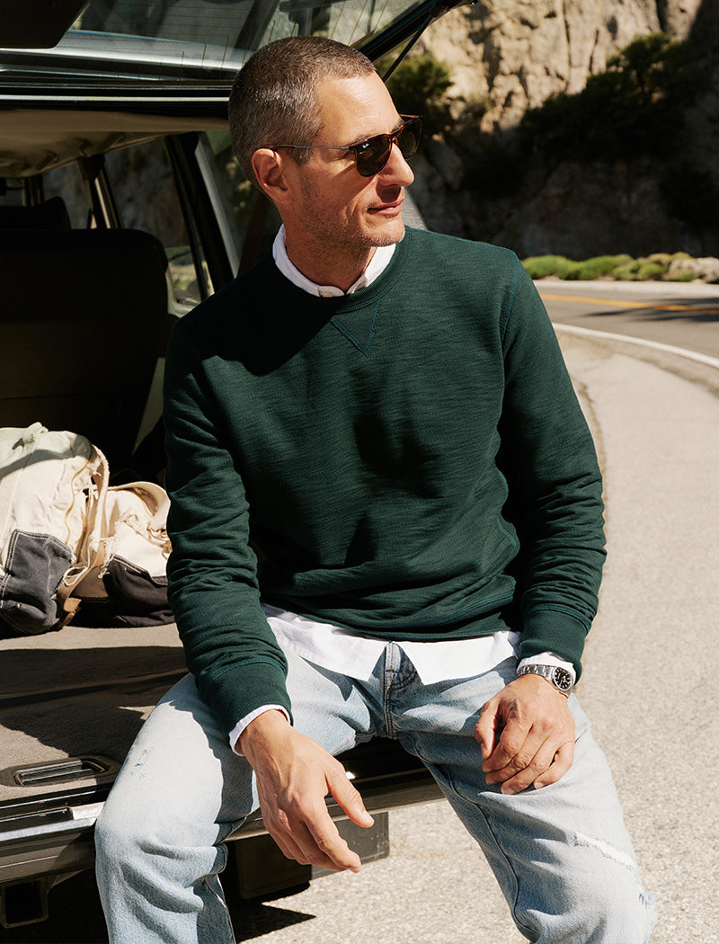 Man sitting by an open car trunk wearing sunglasses and a green sweater.