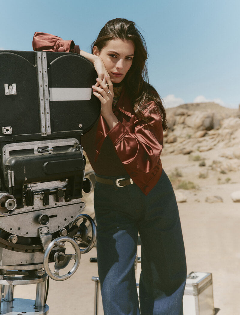 Woman in a red blouse and blue jeans standing next to a vintage movie camera in a desert setting.