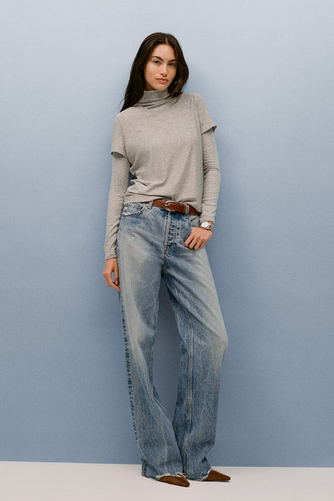 Woman wearing a gray turtleneck sweater and blue jeans against a light blue background